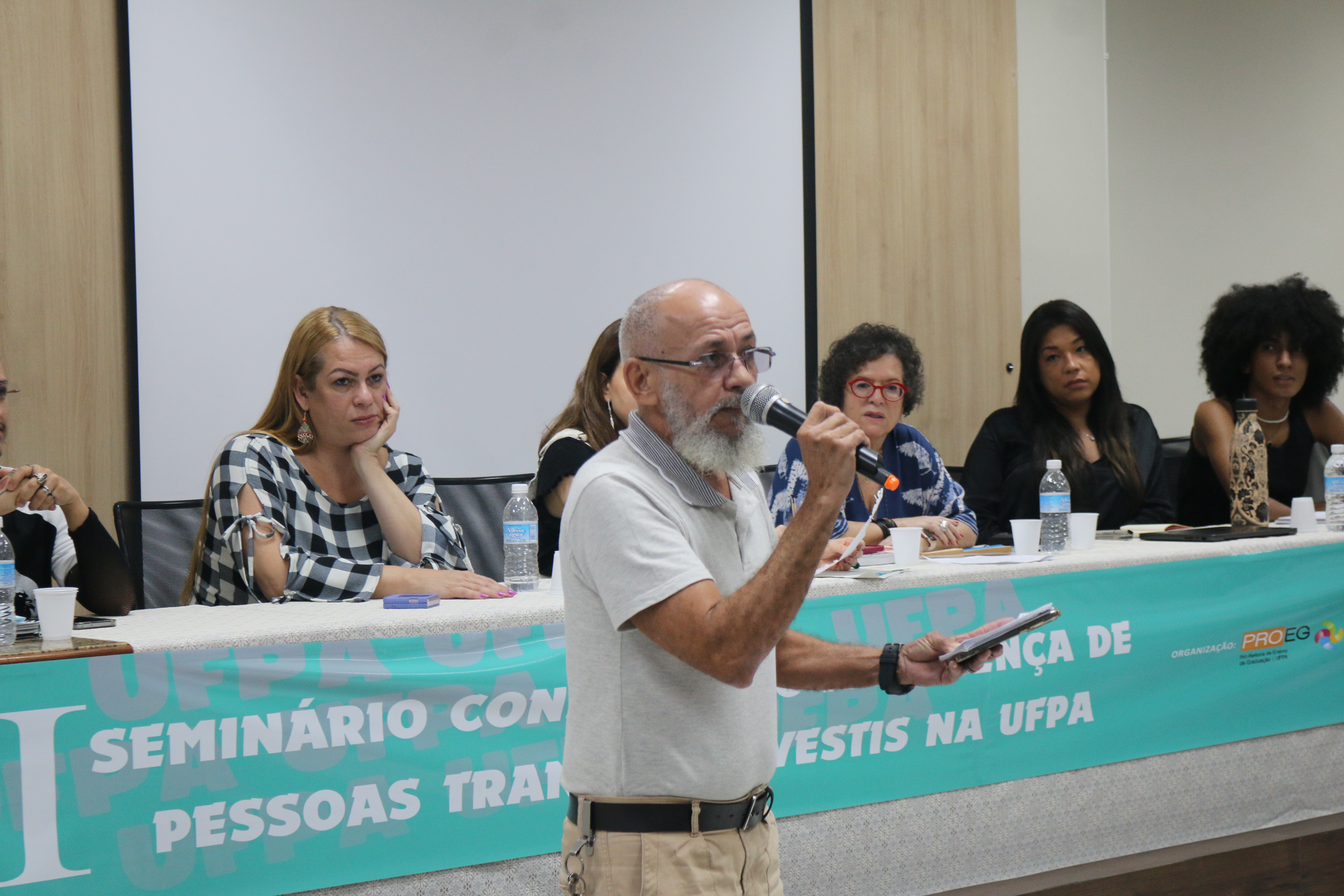 Card informativo sobre o Seminário Construindo a Presença de Pessoas Trans e Travesti na UFPA. A imagem é de um homem idoso à frente do auditório segurando um microfone enquanto fala. Ele é calvo, tem barba branca, usa óculos com lentes quadradas e alça preta. Ele usa camisa cinza, calça cáqui e cinto preto e fala para várias pessoas sentadas à frente dele. Atrás dele, estão cinco mulheres mais um homem do lado esquerdo que quase não aparece. Estão sentados atrás de uma mesa. Na frente dela, há um banner azul claro com  letras brancas, onde se lê: Primeiro Seminário Construindo a Presença de Pessoas Trans e Travesti na UFPA. Ao lado, as logos da UFPA, PROEG, ADIS e ADISTTRAVE.